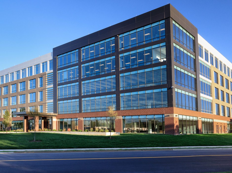 An office building with a brick first level, and black, brown and white panels on the other levels. Around the building is sidewalks and grass.