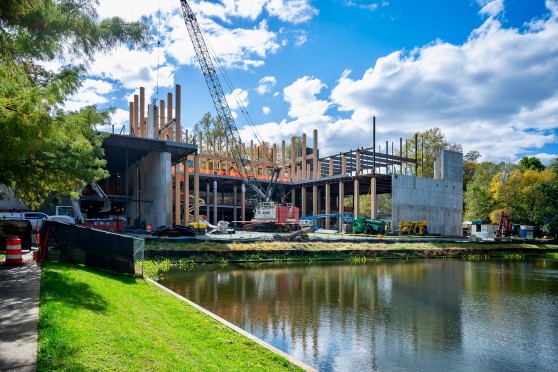 A crane sits in the middle of a concrete building with wooden colums. A lake is in front of the site, and many other machines and building materials are seen around the job.