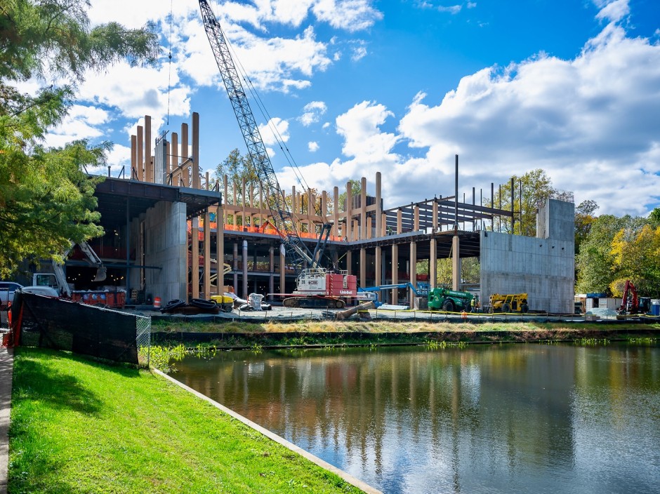 A crane sits in the middle of a concrete building with wooden colums. A lake is in front of the site, and many other machines and building materials are seen around the job.