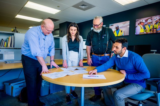 4 people stand around a circular table with construction documents on it.
