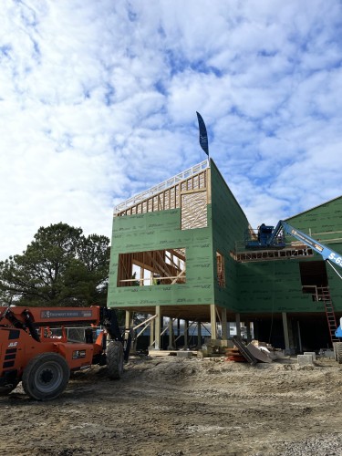 A painted white beam is seen on top of a wooden structure covered in green building material. Large machinery is seen on either side of the building.