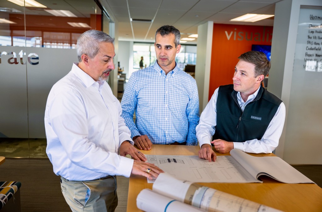 Three men in business attire stand around construction documents and talk.