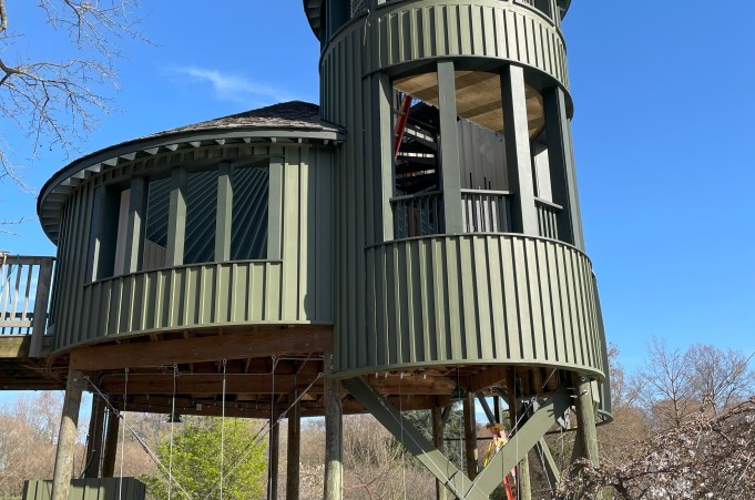 A green circular treehouse with a cone shaper roof is in front of a blue sky