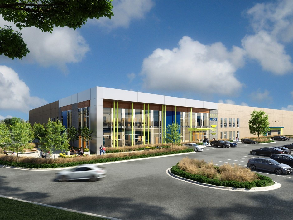 Modern two-story factory building with glass facade and blue and green accents under a clear blue sky. Cars are in the parking lot that sits in front of the building.