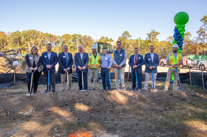 10 people hold shovels in the ground and smile for the camera at a groundbreaking. Behind them is a construction site.